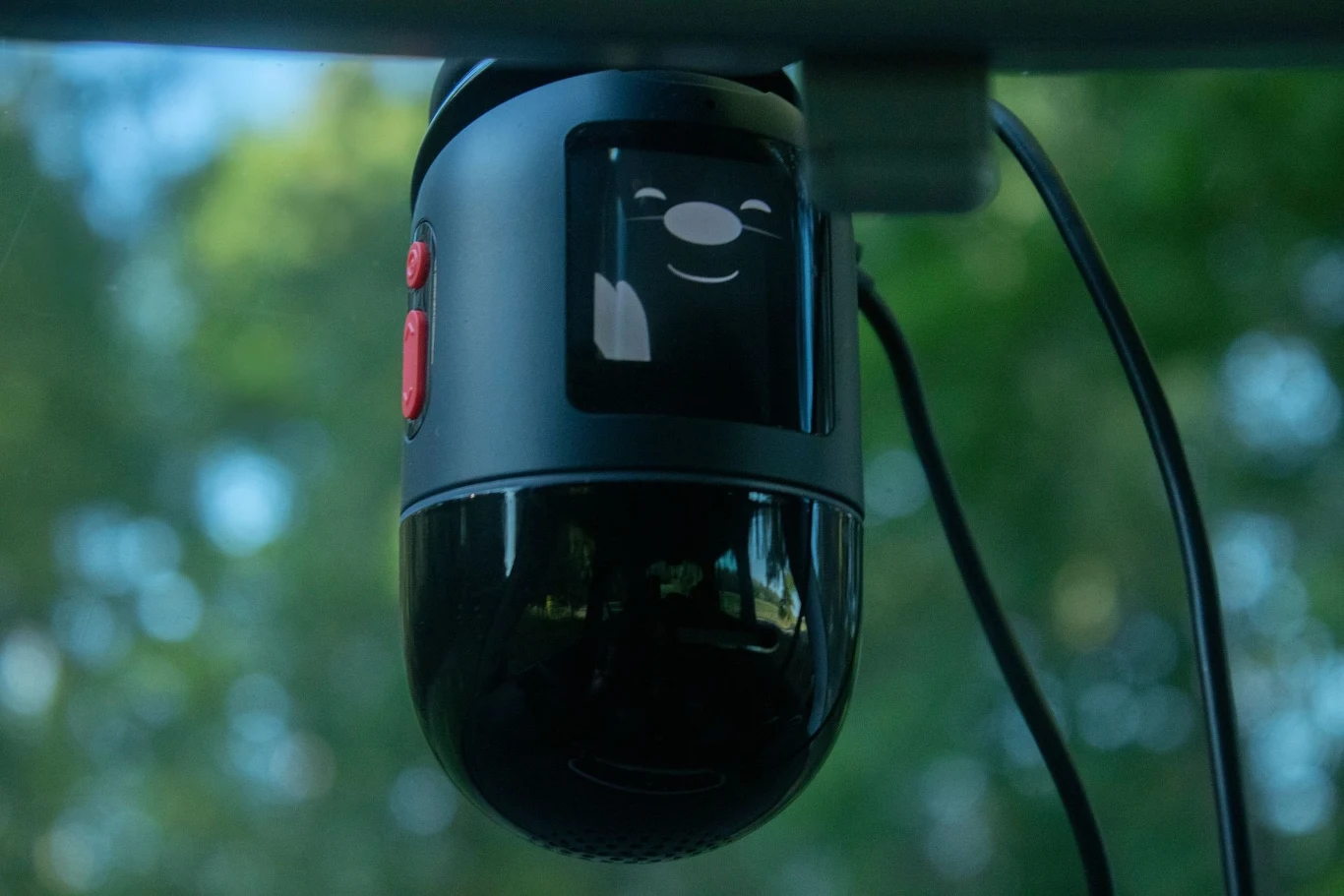 Czarna kamera samochodowa z ekranem wyświetlającym animowaną uśmiechniętą buzię, zamontowana na przedniej szybie pojazdu, z widocznymi przyciskami po bokach i przewodem zasilającym.