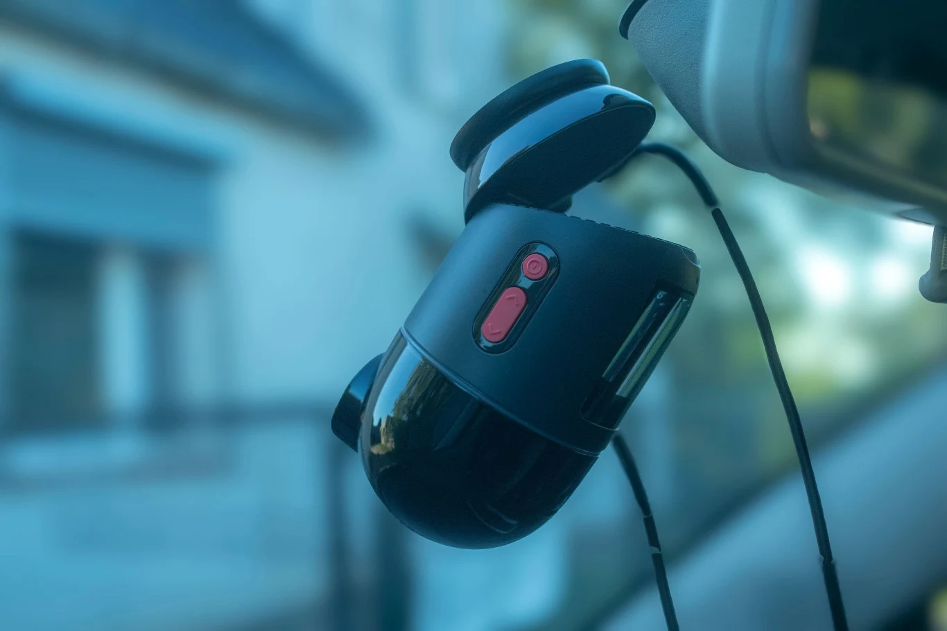 Nowoczesna kamera samochodowa przymocowana do przedniej szyby pojazdu, widoczne wyraźne przyciski funkcyjne i przewód zasilający, w tle zamazany budynek.
