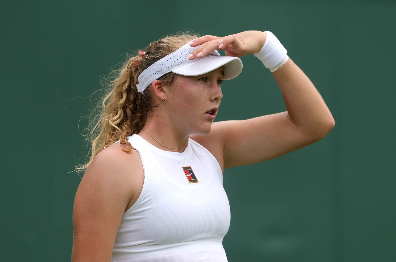 Mirra Andriejewa, Wimbledon 2025 Młoda tenisistka w białym stroju i z daszkiem na głowie, z ręką uniesioną do czoła, stoi na tle zielonej ściany kortu.