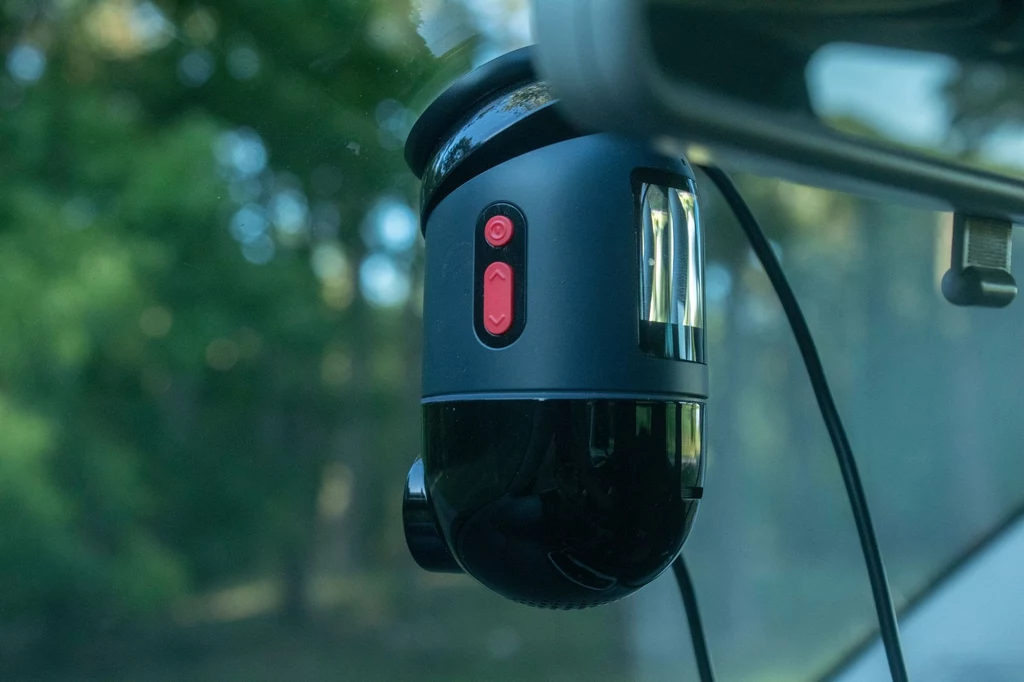 Nie wszystkie funkcje 70mai 4K Omni są dostępne w podstawowym zestawie. Kamera samochodowa zamontowana na przedniej szybie pojazdu, widoczny czerwony przycisk i kabel zasilający, w tle rozmyte drzewa.