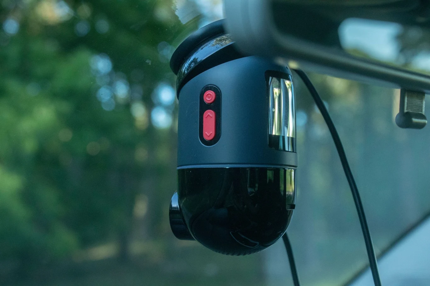 Kamera samochodowa zamontowana na przedniej szybie pojazdu, widoczny czerwony przycisk i kabel zasilający, w tle rozmyte drzewa.
