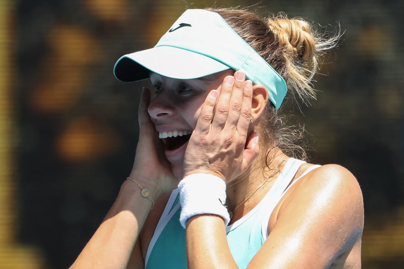 Kobieta w stroju sportowym i czapce z daszkiem wyraża silne emocje, szeroko otwierając usta i trzymając ręce na twarzy, skóra błyszczy od potu, w tle rozmazane, ciemne barwy.