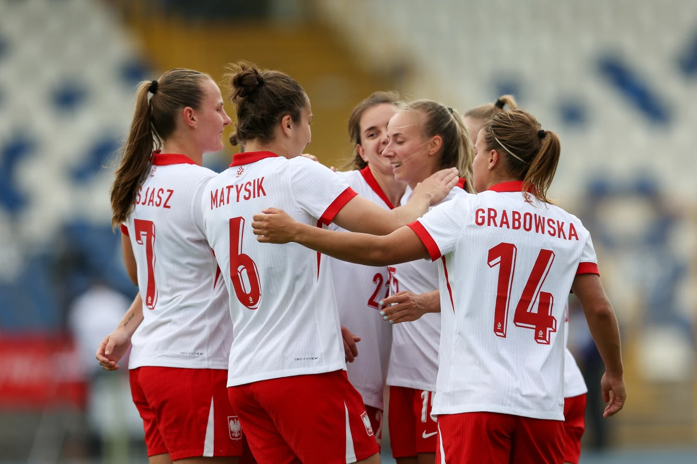 Reprezentantki Polski Grupa piłkarek w biało-czerwonych strojach z numerami i nazwiskami świętuje razem, obejmując się na tle stadionu sportowego.
