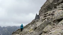 Osoba w niebieskiej kurtce wędruje po kamienistej, górskiej ścieżce, obok skalistego zbocza z rozpiętym namiotem na tle pochmurnego nieba i gór w oddali.