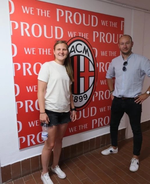 Małgorzata Mesjasz (AC Milan) i Dawid Płaczkiewicz Dwie osoby stoją przy czerwonej ścianie z dużym logo klubu piłkarskiego AC Milan oraz powtarzającym się napisem 'We the proud'. Kobieta ubrana w białą koszulkę i czarne spodenki oraz mężczyzna w jasnej koszuli i ciemnych spodniach pozują do zdjęcia.