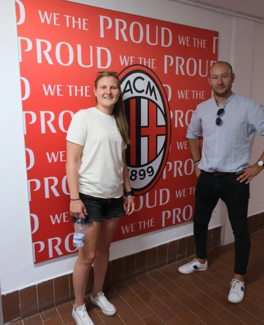 Małgorzata Mesjasz (AC Milan) i Dawid Płaczkiewicz Dwie osoby stoją przy czerwonej ścianie z dużym logo klubu piłkarskiego AC Milan oraz powtarzającym się napisem 'We the proud'. Kobieta ubrana w białą koszulkę i czarne spodenki oraz mężczyzna w jasnej koszuli i ciemnych spodniach pozują do zdjęcia.