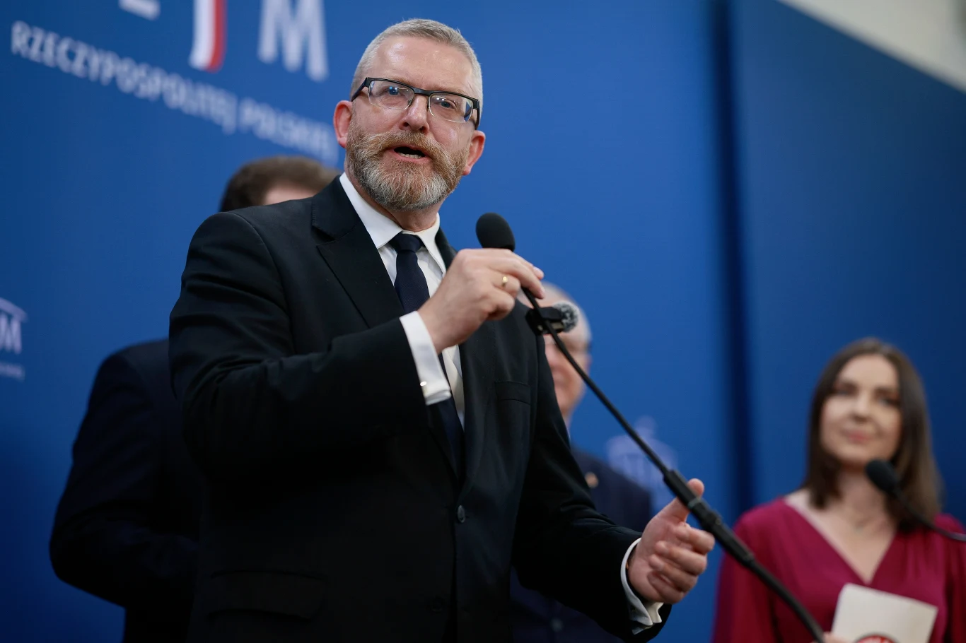 Prokuratura podsumowała czynności z udziałem Grzegorza Brauna Mężczyzna w garniturze i okularach przemawia do mikrofonu na tle niebieskiego baneru z logo oraz napisem sugerującym oficjalne wydarzenie, w tle kilka innych osób, jedna z kobiet trzyma dokumenty.