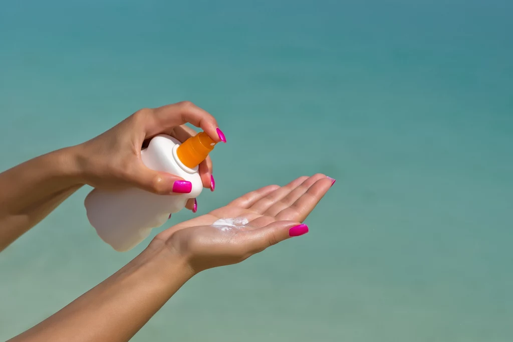 Ochrona przed słońcem jest ważna nie tylko przed opalaniem Ręka z różowym manicure trzyma butelkę kremu z filtrem i wyciska go na drugą dłoń, w tle rozmazana powierzchnia wodna w jasnym, turkusowym kolorze.