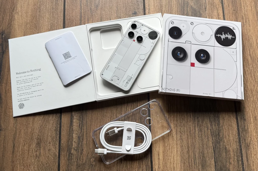 Zawartość opakowania z Nothing Phone 3. Otwarty zestaw sprzedażowy smartfona Nothing Phone (2), stylizowany w kolorze białym z przezroczystymi elementami, zawiera telefon, plastikowe przezroczyste etui, przewód USB oraz instrukcję w eleganckim pudełku na drewnianej powierzchni.
