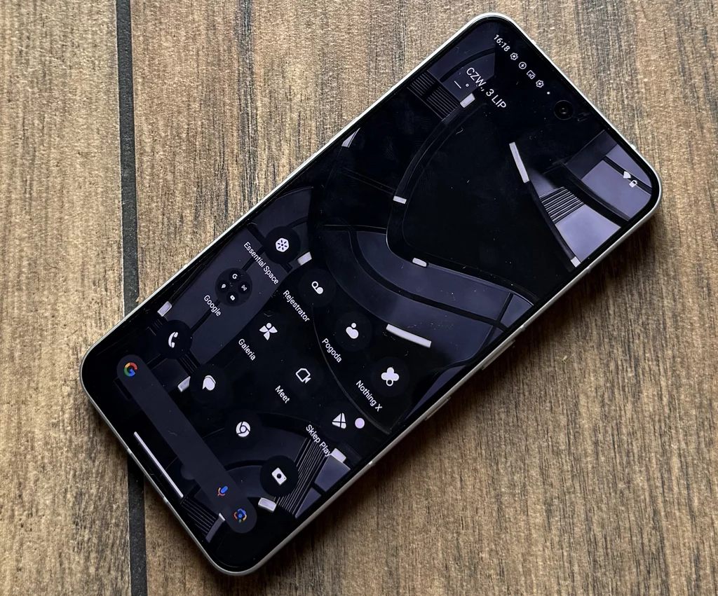Nothing OS to oprogramowanie smartfona Nothing Phone 3. Nowoczesny smartfon leżący na drewnianej powierzchni, z wyświetlonym na ekranie menu w trybie ciemnym oraz ikonami popularnych aplikacji i narzędzi systemowych.