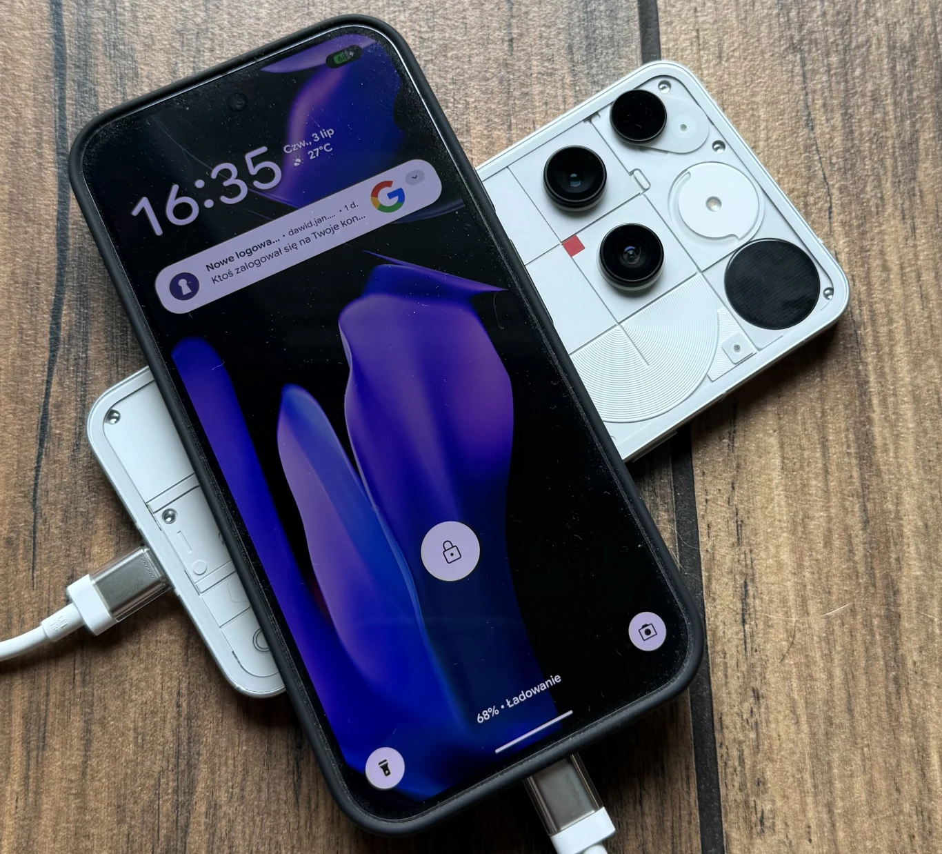 Dwa smartfony leżą na drewnianej powierzchni, oba są podłączone do ładowania przewodowego, na pierwszym planie widoczny ekran urządzenia z powiadomieniami i godzina, w tle widać drugi telefon z charakterystycznym przezroczystym panelem i trzema aparatami.