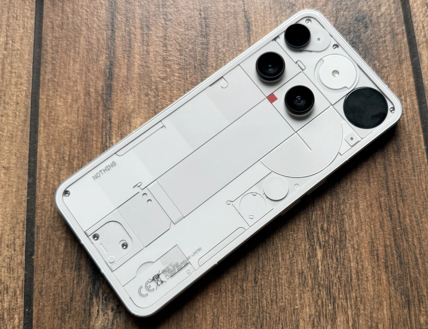 Nowoczesny smartfon marki Nothing wykończony przezroczystą obudową ukazującą elementy wewnętrzne, położony na drewnianej powierzchni; urządzenie wyposażone w trzy aparaty tylne oraz wyraźne detale konstrukcyjne.