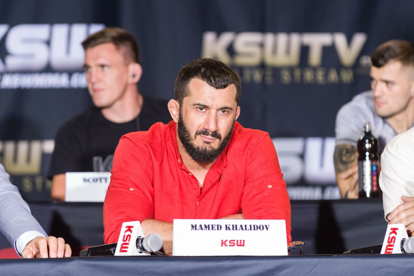 Mężczyzna z brodą w czerwonej koszuli siedzi przy stole konferencyjnym z mikrofonami i tabliczką z imieniem i nazwiskiem. W tle logo KSW TV i inni uczestnicy konferencji.