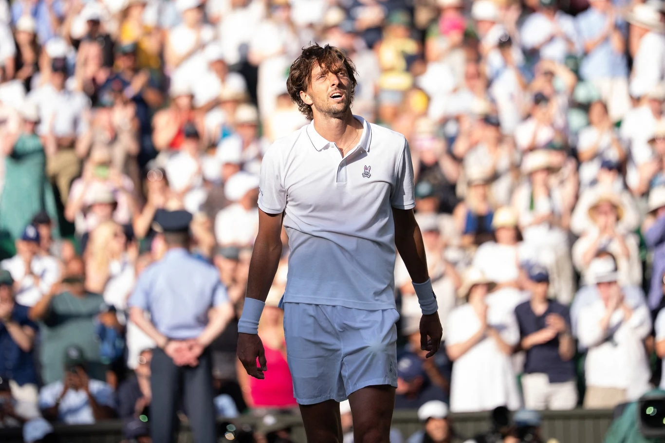 Arthur Rinderknech na Wimbledonie Tenisista w białym stroju stoi na korcie podczas meczu, wokół tłum widzów na trybunach, intensywne światło podkreśla mocne emocje sportowca.