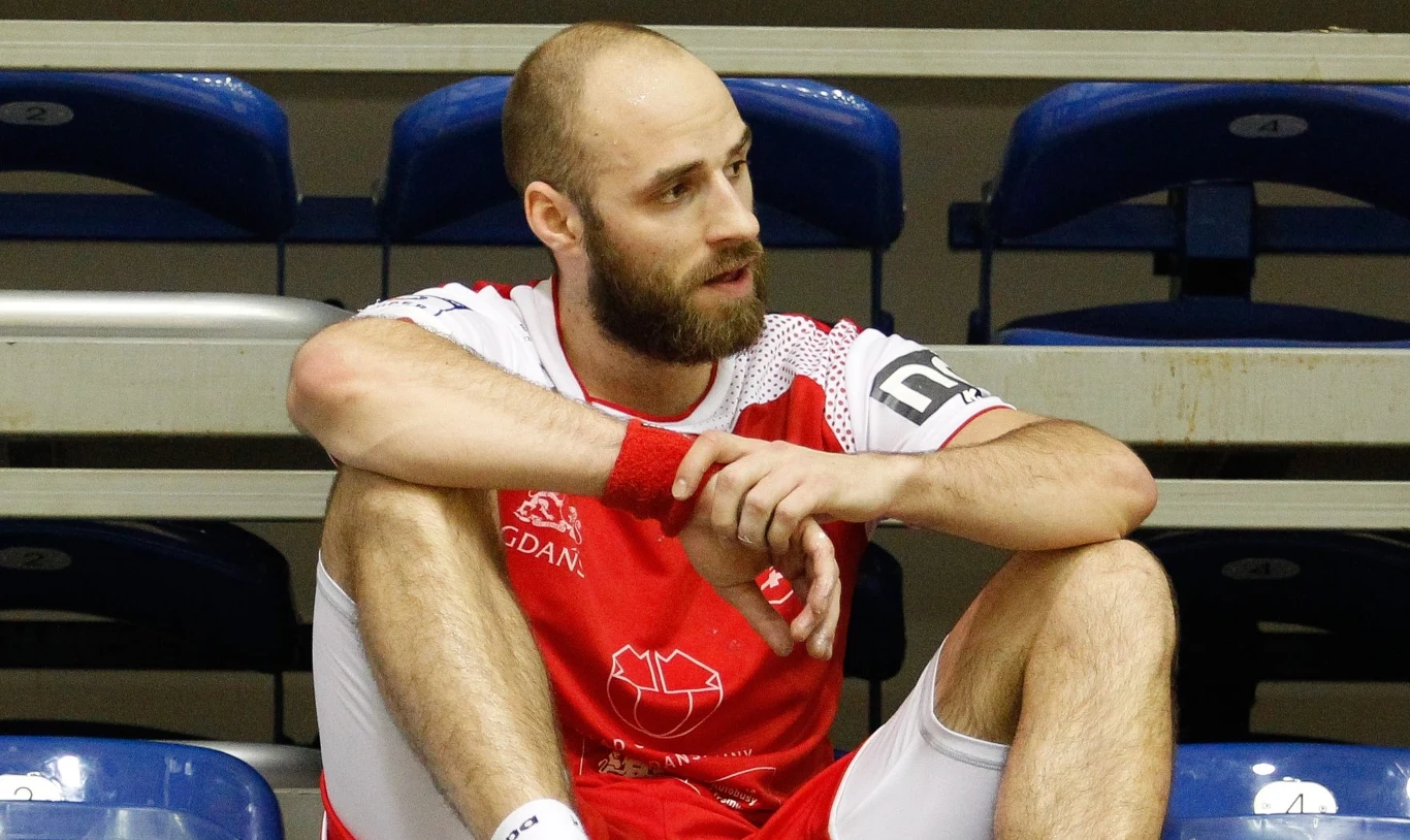 Sportowiec w biało-czerwonym stroju siedzący na stopniach trybun, wygląda na zamyślonego lub zmęczonego, na pierwszym planie widoczne umięśnione nogi i ręce, w tle niebieskie plastikowe krzesełka.