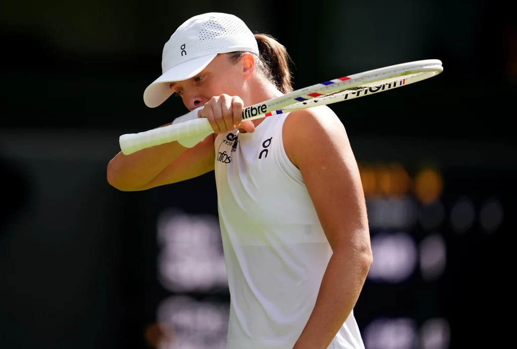 Iga Świątek Tenisistka ubrana w biały strój sportowy z białą czapką odwraca głowę i opiera rakietę tenisową o ramię na tle rozmytego kortu.