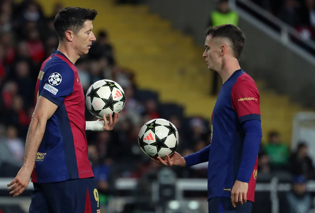 Robert Lewandowski i Marc Casado Dwóch piłkarzy w strojach klubowych na boisku trzymających piłki do gry, skupieni na rozmowie lub przygotowaniach do rozegrania fragmentu meczu, w tle rozmyty tłum widzów na trybunach.