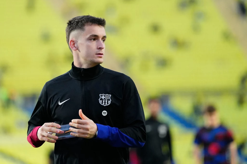 Marc Casado Młody mężczyzna w sportowej kurtce z herbem FC Barcelony stoi na tle stadionu, trzymając w dłoni butelkę wody, w tle rozmyci inni zawodnicy i żółte trybuny.