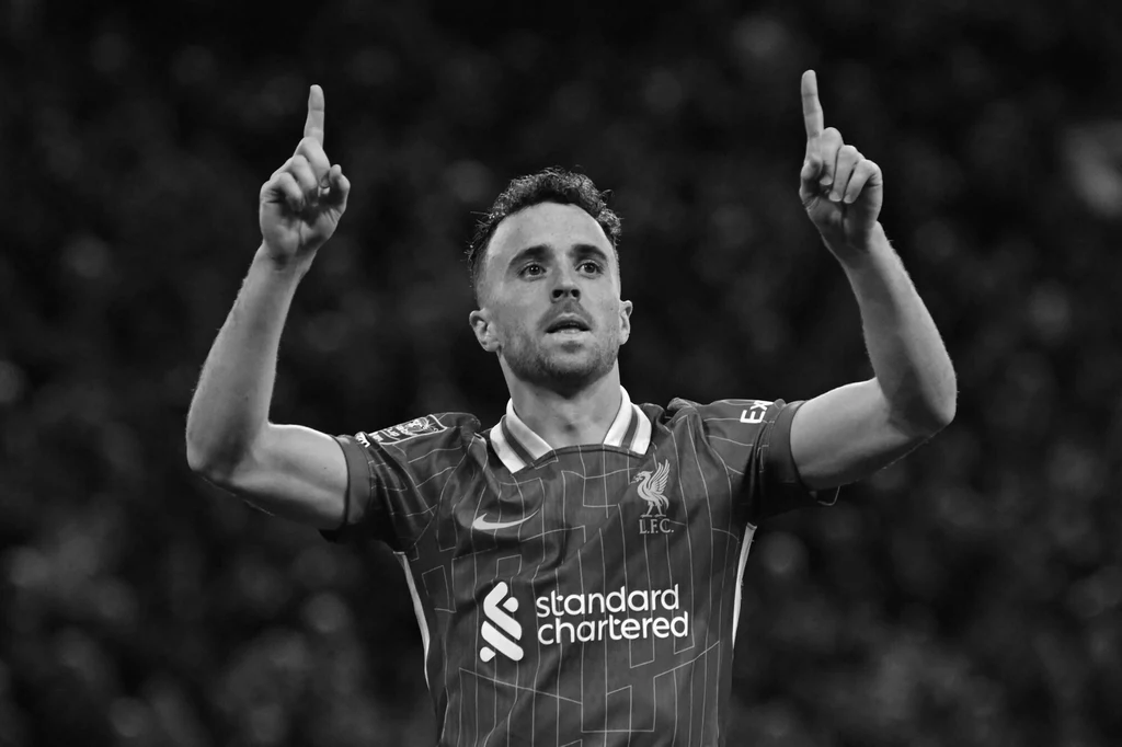 Diogo Jota Piłkarz w koszulce Liverpool FC z uniesionymi obiema rękami i palcami wskazującymi do góry na tle rozmytej publiczności stadionu.