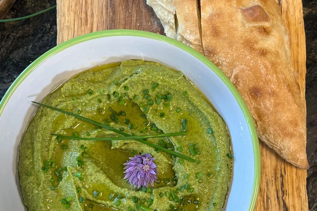 Pasta z bobu i awokado Zielony dip udekorowany szczypiorkiem, oliwą oraz fioletowym kwiatem szczypiorku w białej misce, obok świeży chleb na drewnianej desce.