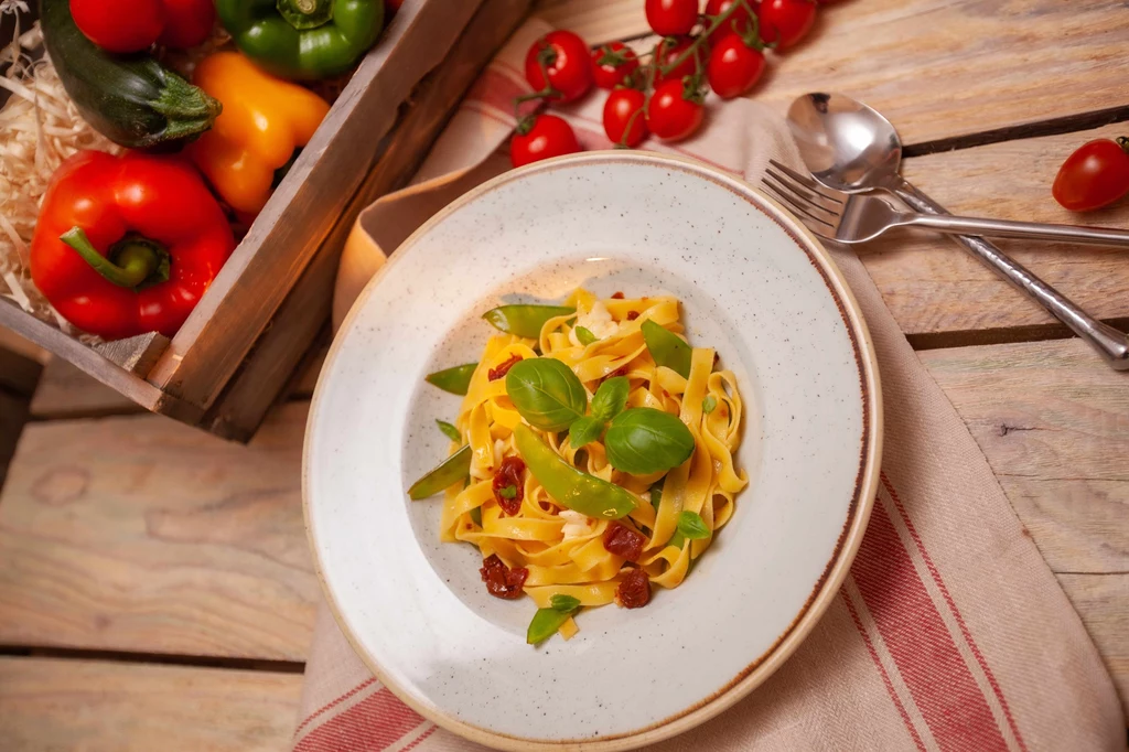 Makaron z suszonymi pomidorami i groszkiem cukrowym Talerz makaronu tagliatelle z suszonymi pomidorami i świeżymi liśćmi bazylii, obok widoczne świeże warzywa – papryka i pomidorki koktajlowe, a całość ustawiona na drewnianym stole przykrytym lnianą serwetką.