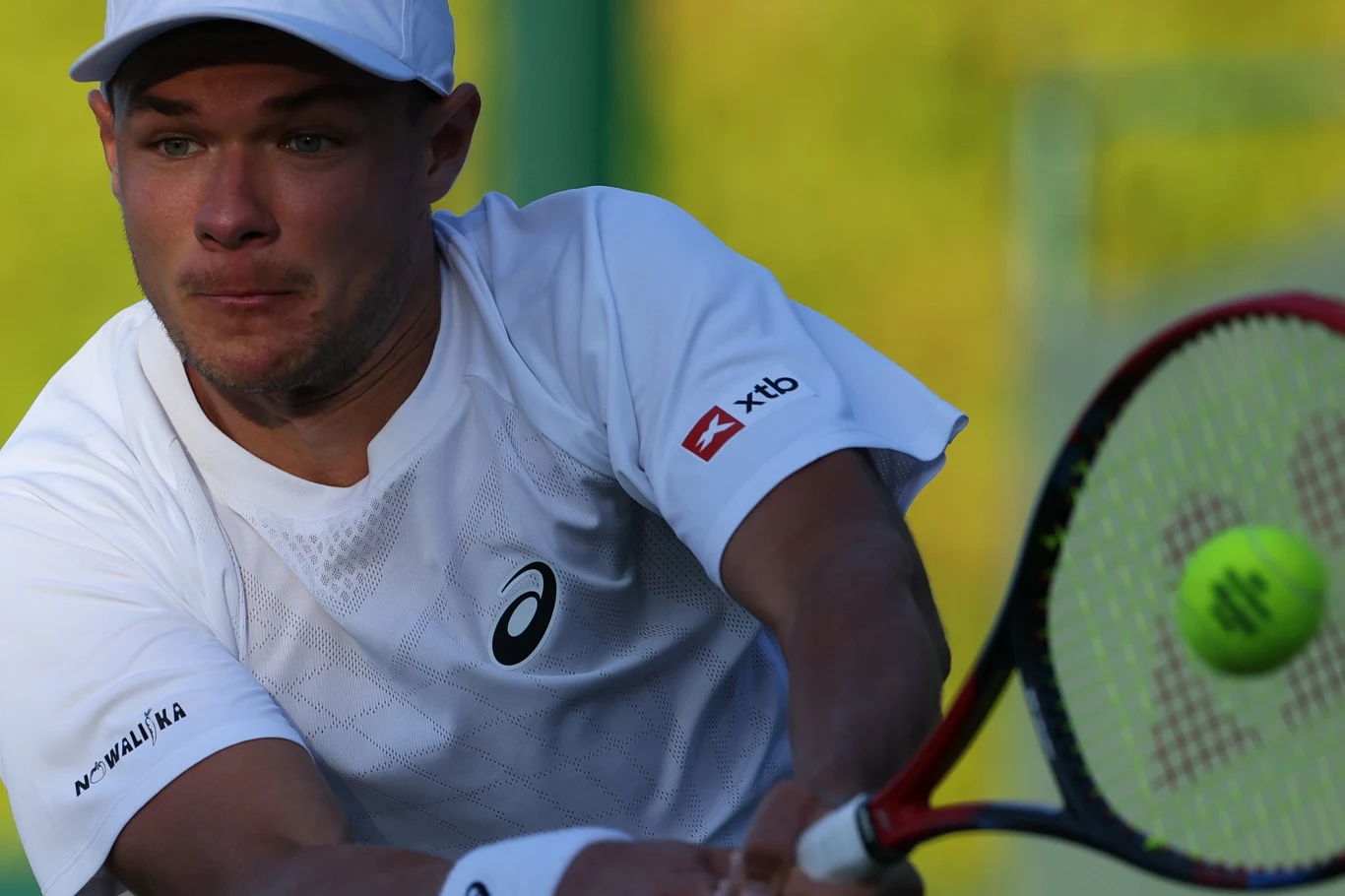 Kamil Majchrzak Zawodnik w białej koszulce i czapce koncentruje się podczas odbicia piłki tenisowej rakietą, tło rozmyte z przewagą zieleni, widoczne logo sponsora na rękawie.