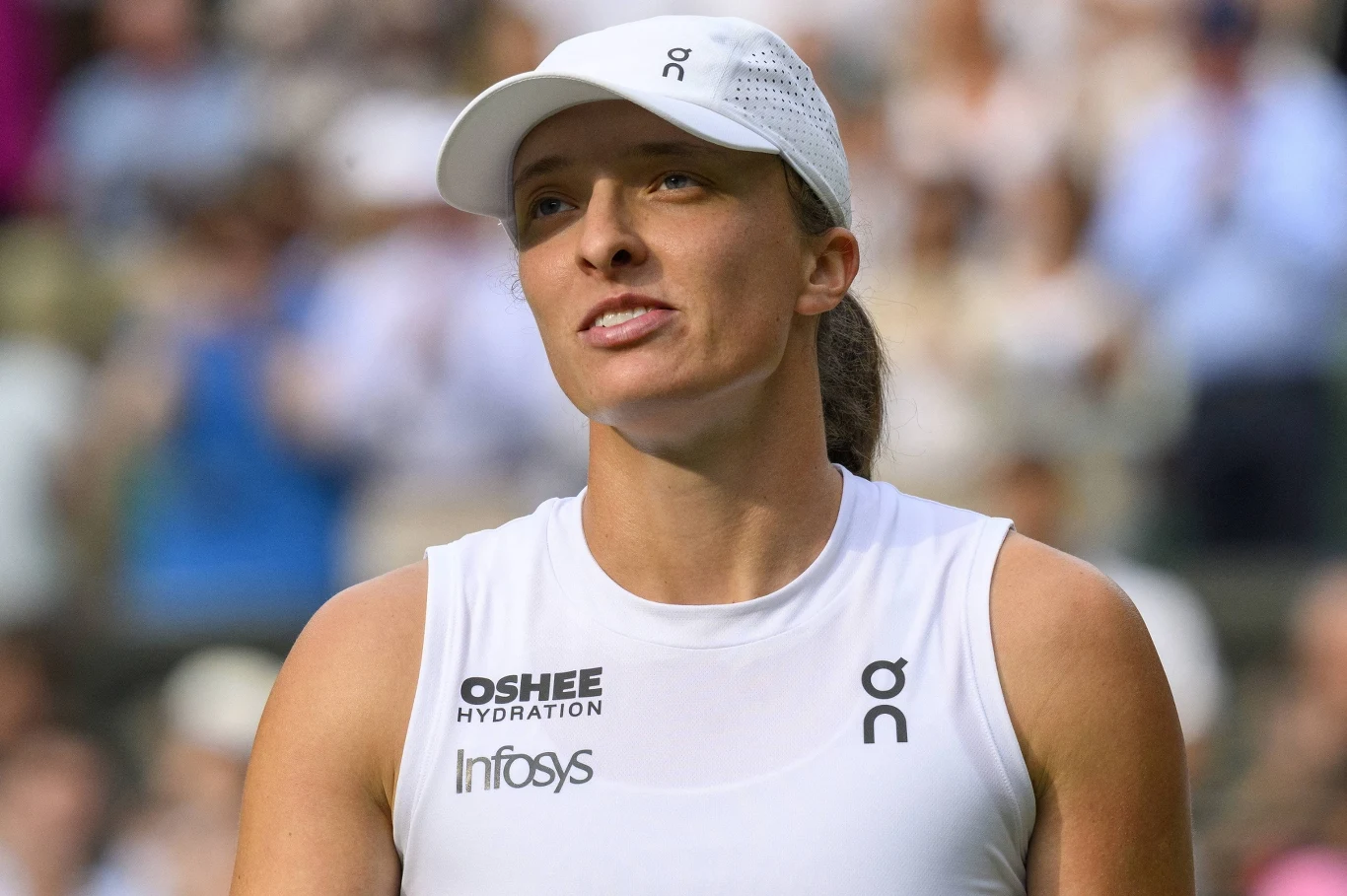 Iga Świątek Tenisistka ubrana w biały sportowy strój z widocznymi logotypami sponsorów i białą czapką z daszkiem, w tle rozmazana publiczność.