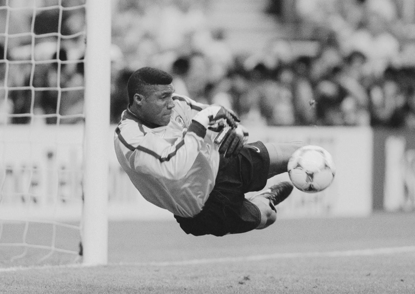 Peter Rufai Bramkarz w sportowym stroju wykonuje dynamiczną paradę, próbując obronić piłkę lecącą w kierunku bramki podczas meczu piłki nożnej, w tle rozmazany tłum widzów.