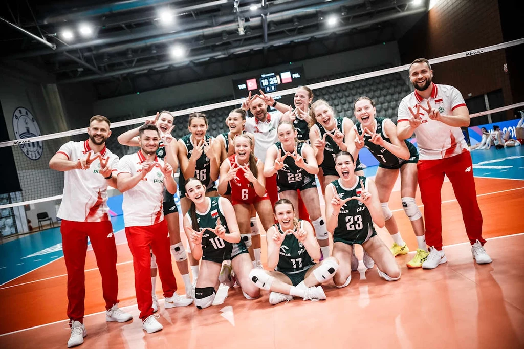 Reprezentacja Polski siatkarek do lat 19 Grupa siatkarek oraz trenerów uśmiecha się, pozując na boisku do siatkówki po zakończonym meczu, wszyscy ubrani w stroje sportowe, wykonując gesty zwycięstwa rękami, w tle widoczna siatka oraz tablica wyników.