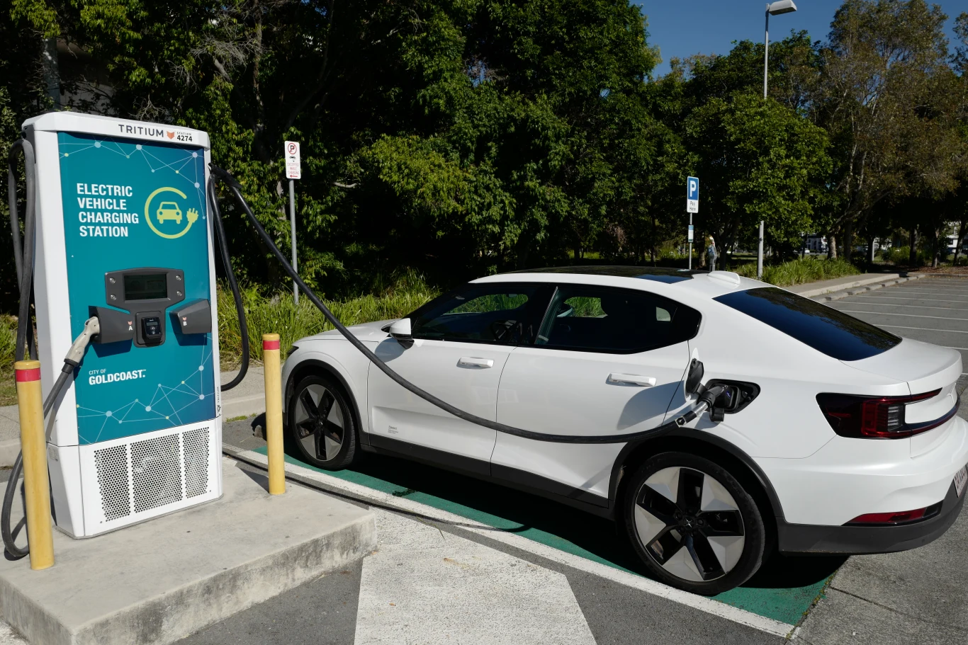 O ile latem spada zasięg samochodów elektrycznych? Biały samochód elektryczny ładowany przy stacji ładowania, otoczony zielenią i stojący na oznaczonym miejscu parkingowym.