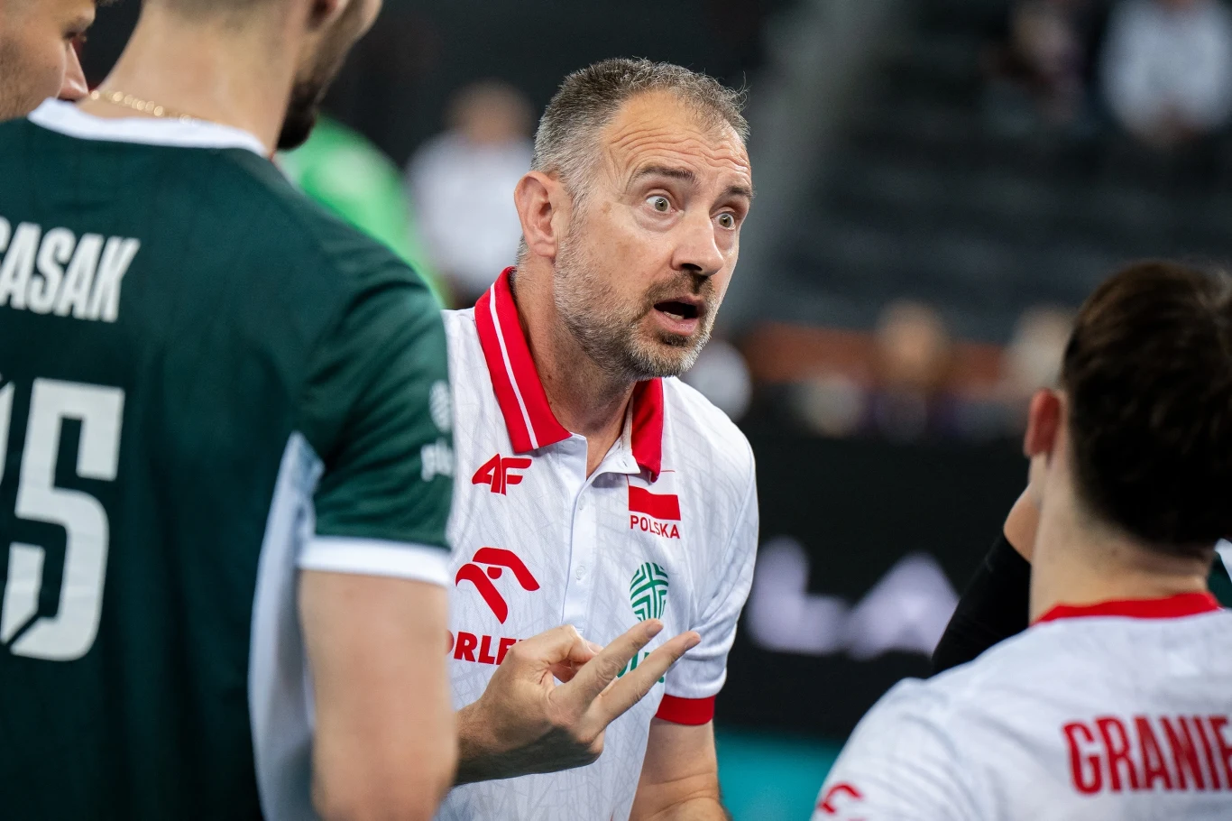 Trener reprezentacji Polski w siatkówce udziela wskazówek zawodnikom podczas meczu, wyrażając silne emocje i aktywnie gestykulując, wokół niego stoją zawodnicy w sportowych strojach z widocznymi nazwiskami na plecach.