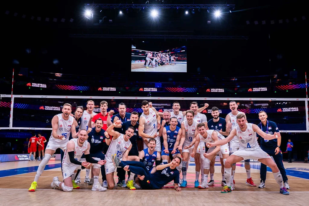 Pavle Perić występuje tego lata w reprezentacji Serbii Grupa siatkarzy pozujących wspólnie na środku boiska, uśmiechnięci i radośni, część ubrana w białe stroje sportowe, część w ciemnych, w tle siatka i rozświetlone trybuny sowie ekran pokazujący powtórkę drużyny.