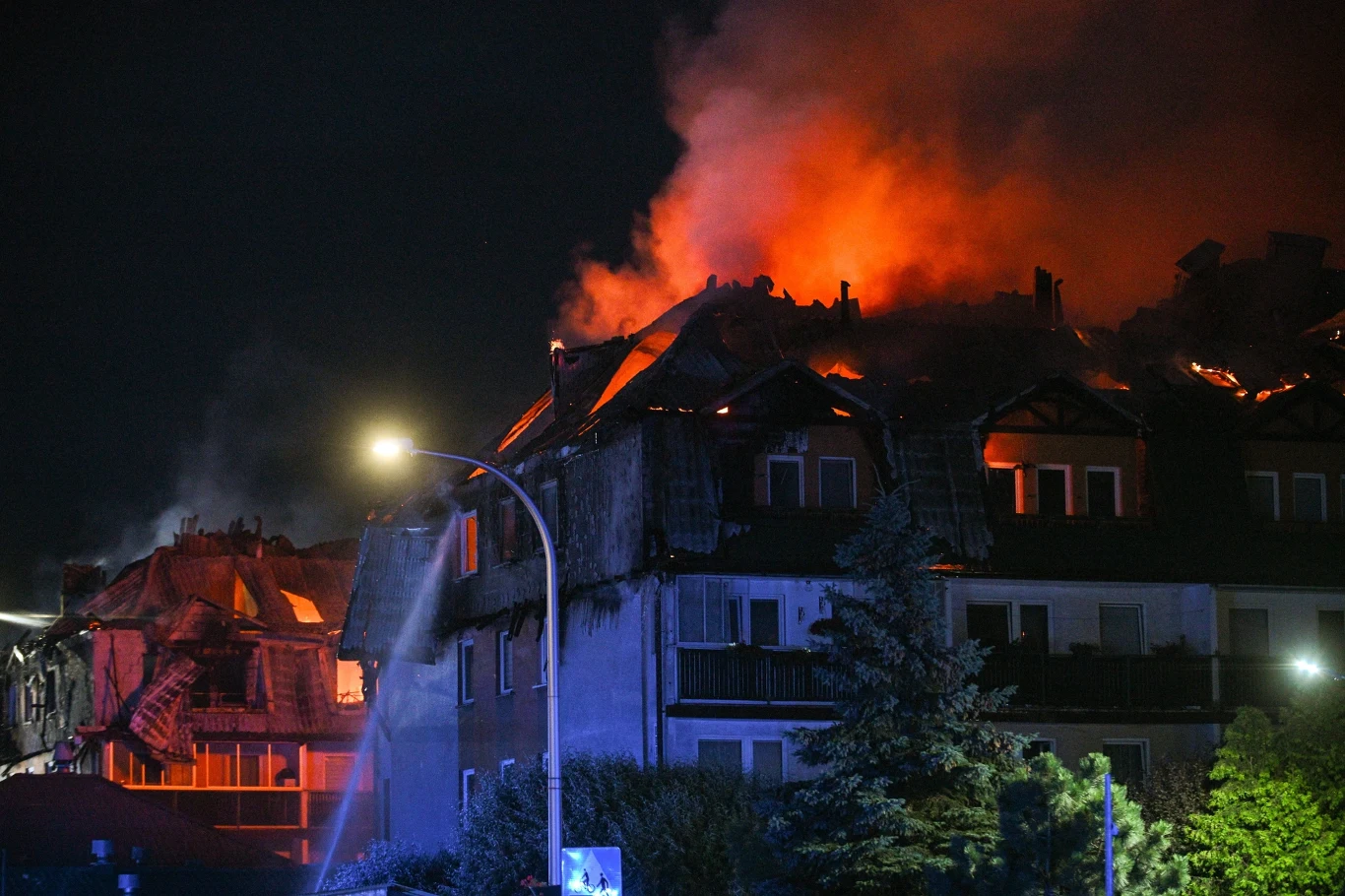 Pożar w Ząbkach Płonący dach budynku mieszkalnego w nocy, intensywne płomienie oraz kłęby dymu unoszące się wysoko nad konstrukcją, na pierwszym planie widoczna latarnia uliczna i drzewa.