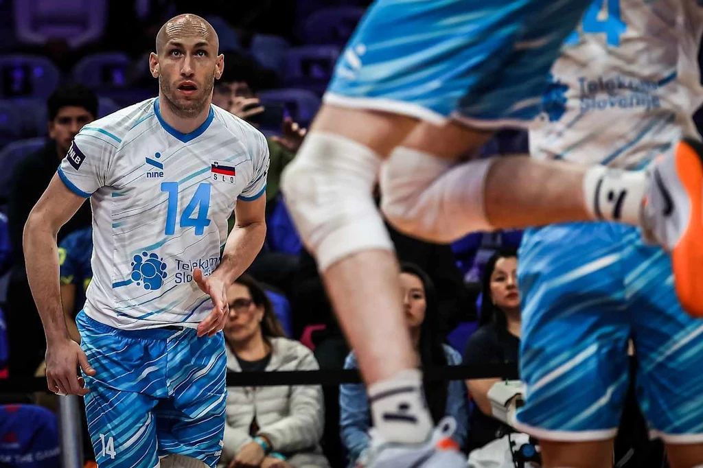 Żiga Stern w barwach reprezentacji Słowenii Siatkarz w niebieskim stroju sportowym z numerem 14 i emblematem reprezentacyjnym skupiony podczas meczu siatkarskiego, na pierwszym planie widoczna noga innego zawodnika w ruchu, w tle publiczność obserwująca rozgrywkę.