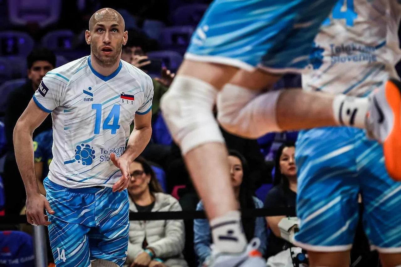 Żiga Stern w barwach reprezentacji Słowenii Siatkarz w niebieskim stroju sportowym z numerem 14 i emblematem reprezentacyjnym skupiony podczas meczu siatkarskiego, na pierwszym planie widoczna noga innego zawodnika w ruchu, w tle publiczność obserwująca rozgrywkę.