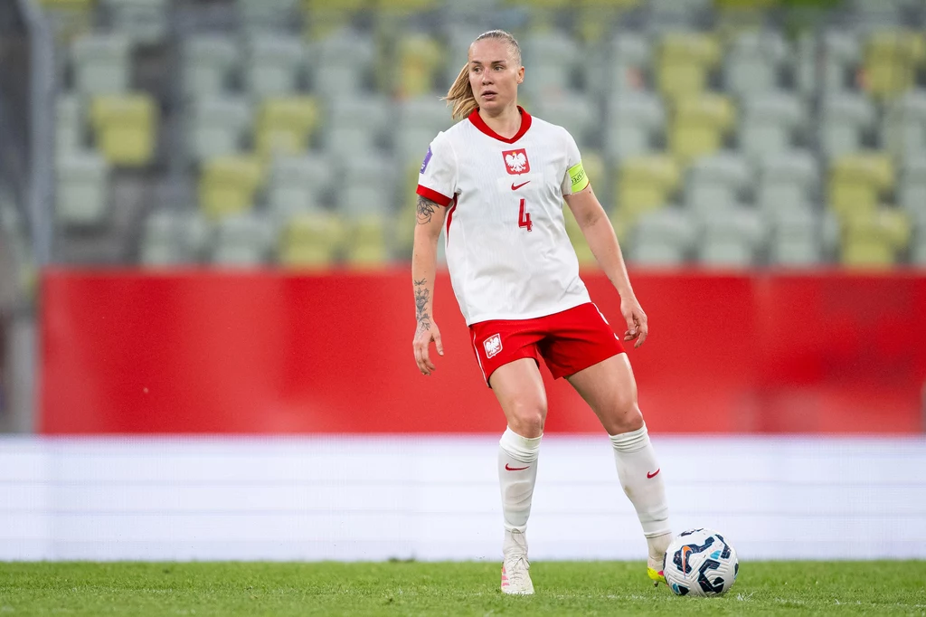 Paulina Dudek w barwach reprezentacji Polski kobiet Zawodniczka reprezentacji Polski w piłce nożnej w stroju meczowym, z opaską kapitańską na ramieniu, kontroluje piłkę na boisku podczas meczu.