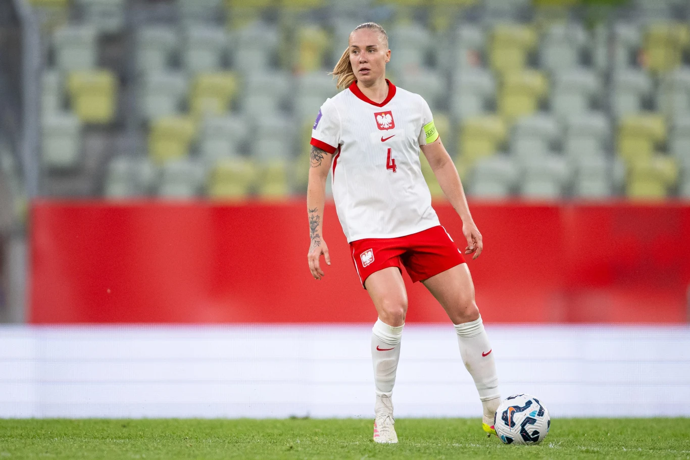 Paulina Dudek w barwach reprezentacji Polski kobiet Zawodniczka reprezentacji Polski w piłce nożnej w stroju meczowym, z opaską kapitańską na ramieniu, kontroluje piłkę na boisku podczas meczu.
