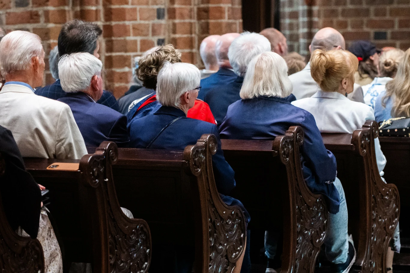 Msza w kościele, zdj. ilustracyjne Grupa starszych osób siedzi w ozdobnych drewnianych ławkach w budynku o ceglanych ścianach, zwróconych plecami do obiektywu, uczestnicząc w wydarzeniu lub uroczystości.