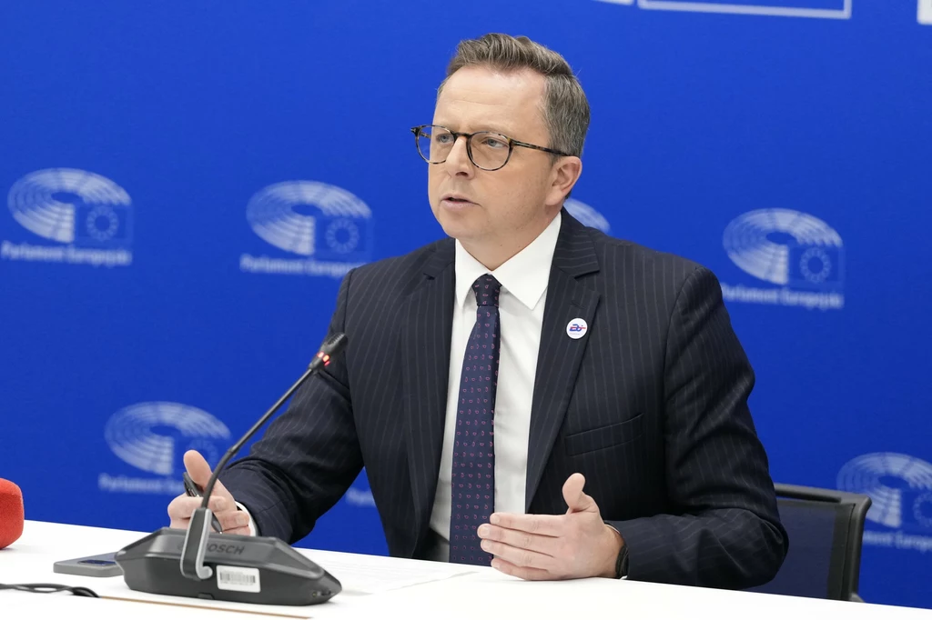 Dariusz Joński Mężczyzna w okularach, garniturze i krawacie siedzi przy stole z mikrofonem, przemawia podczas konferencji prasowej na tle logotypów Parlamentu Europejskiego na niebieskiej ścianie.
