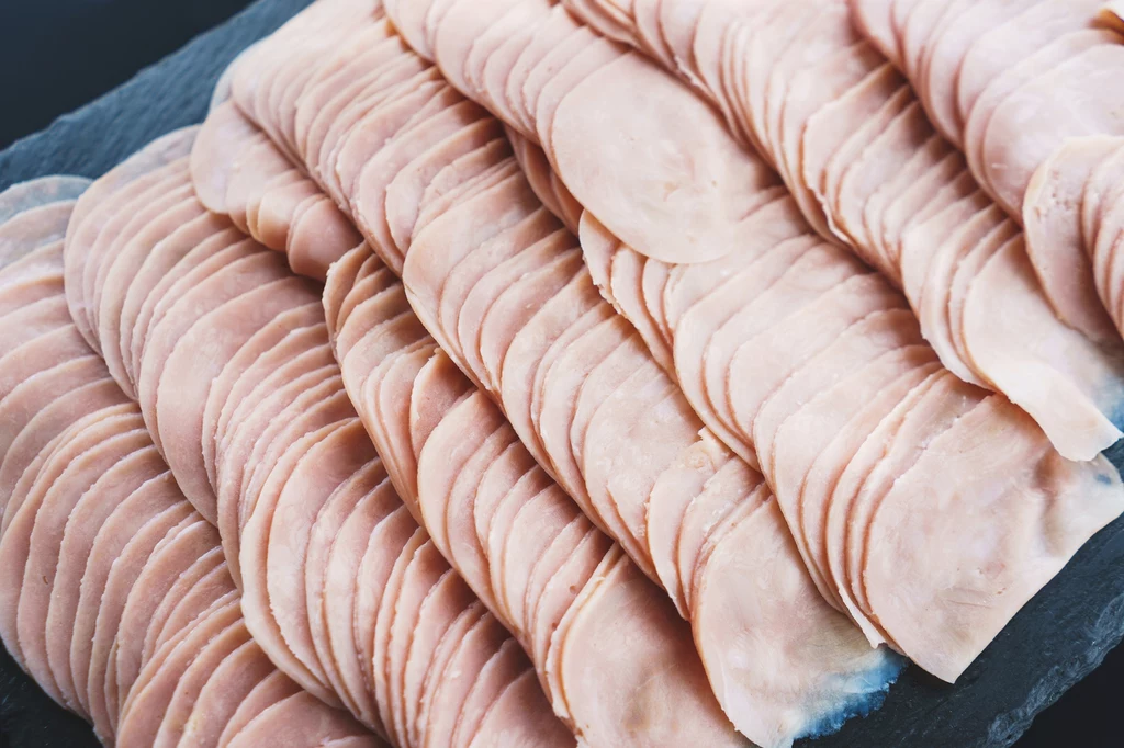 Szynki o zwartej i jednolitej strukturze cechują się większą tendencją do powstawania tęczowej poświaty Cienko pokrojone plastry szynki poukładane w równych rzędach na ciemnym tle.
