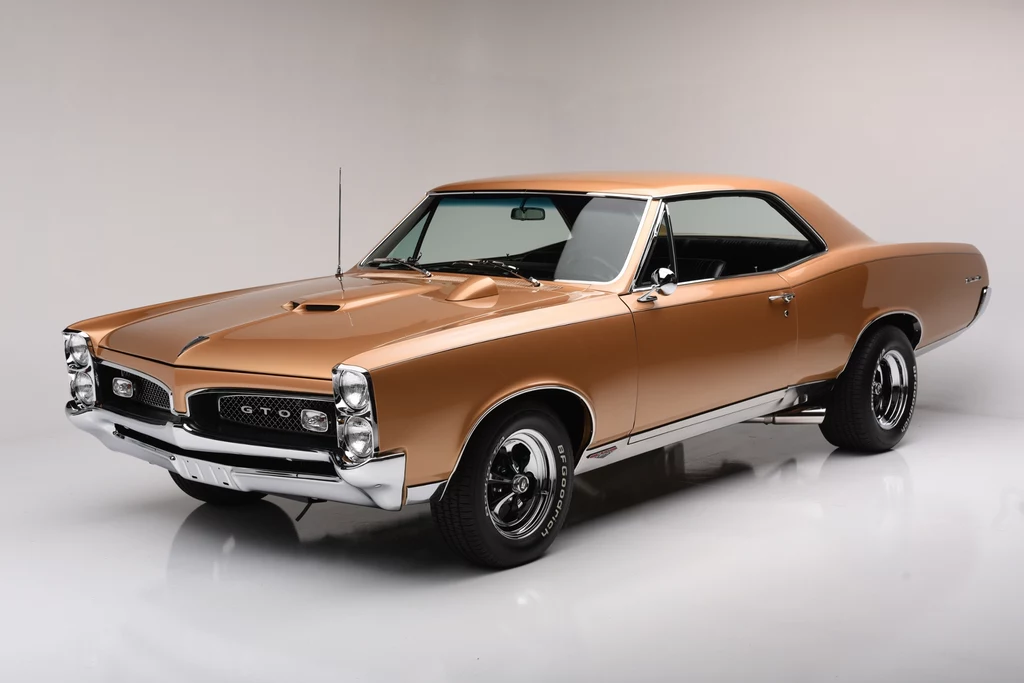 Złoty klasyczny samochód Pontiac GTO z lat 60., z czarnym dachem i wyrazistymi, chromowanymi detalami, ustawiony na białym tle studyjnym. Wyraźnie widoczny grill z logo oraz masywne koła.