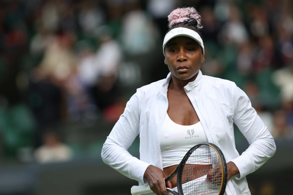 Venus Williams Tenisistka w białym stroju sportowym trzymająca rakietę na korcie podczas meczu, w tle rozmyta publiczność.