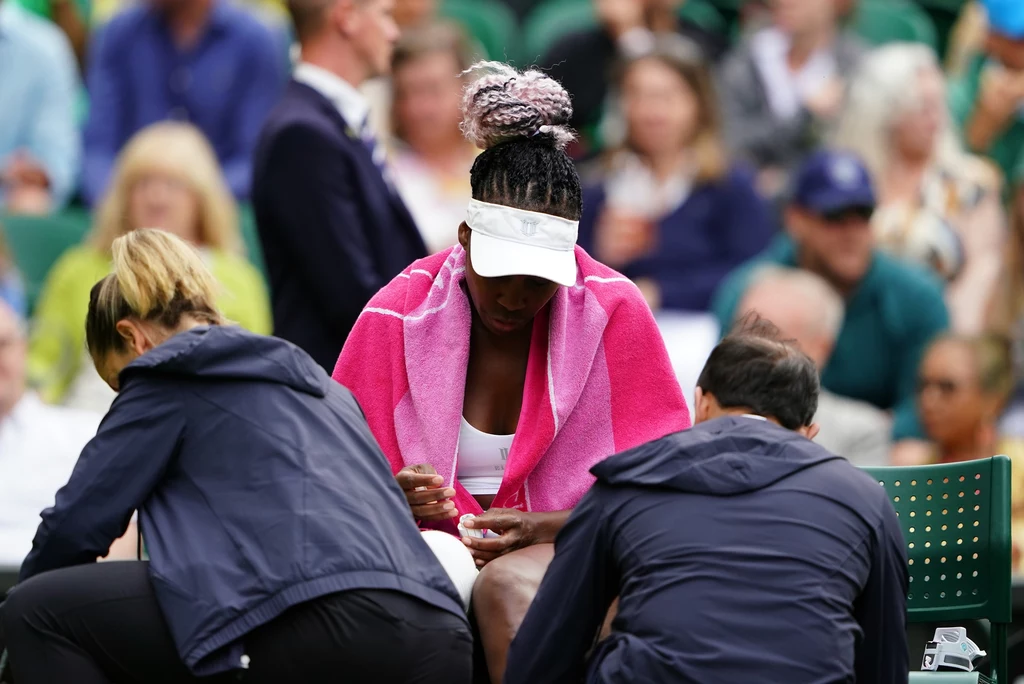 Venus Williams Zawodniczka tenisowa siedzi na ławce owinięta różowym ręcznikiem, a dwóch pracowników medycznych udziela jej pomocy, podczas gdy w tle widoczna jest publiczność.