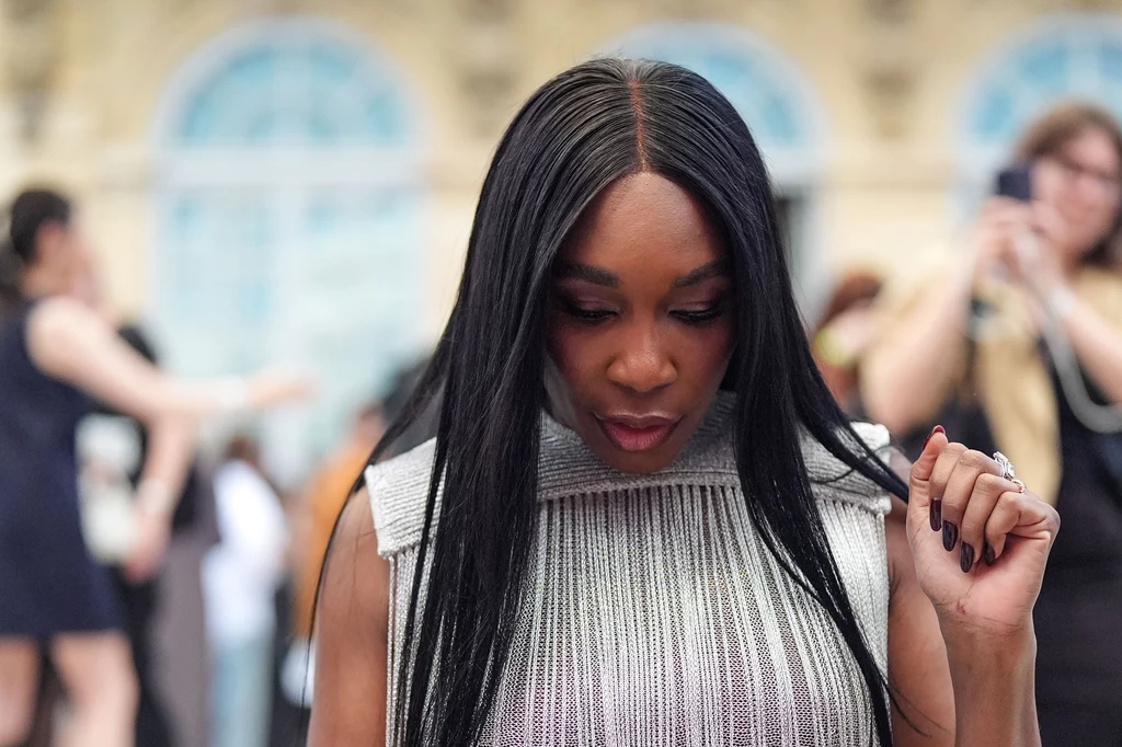 Venus Williams Kobieta o długich czarnych prostych włosach ubrana w jasny, elegancki top, pochylona lekko do przodu, w tle rozmyte sylwetki innych ludzi oraz jasne budynki z dużymi oknami.