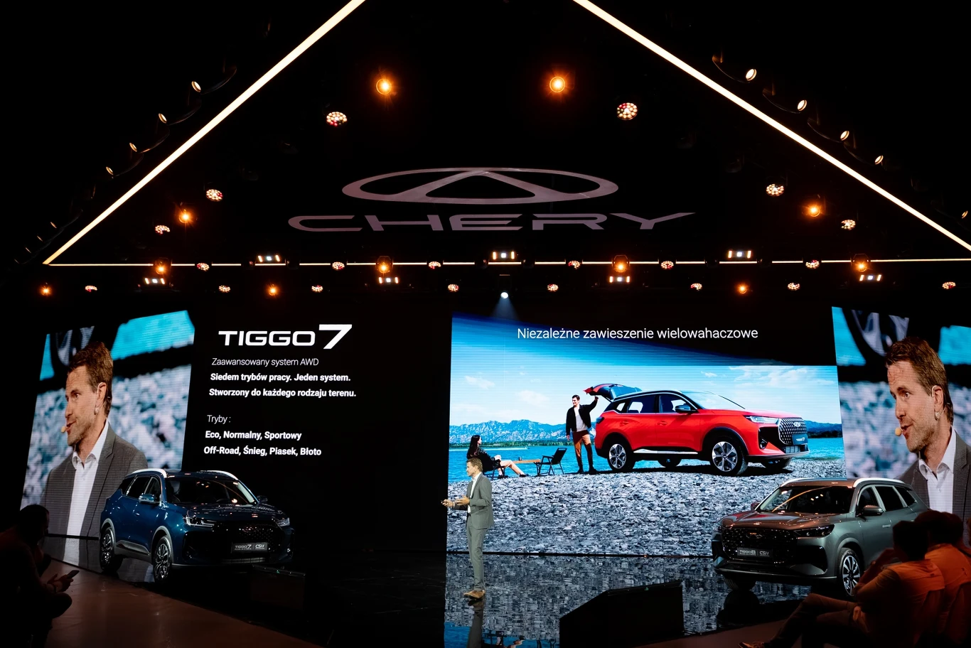 Prezentacja nowego modelu samochodu Chery Tiggo7 na scenie z dużym podświetlonym logo marki, osobą prowadzącą oraz dużymi ekranami wyświetlającymi zdjęcia auta i informacje techniczne.