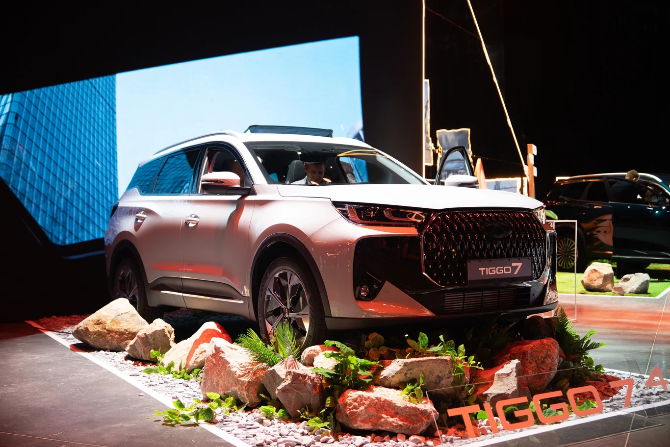Nowoczesny SUV marki Tiggo 7 ustawiony na platformie dekoracyjnej otoczonej skałami i roślinnością, w tle fragment szklanego biurowca oraz elementy ekspozycji targowej.