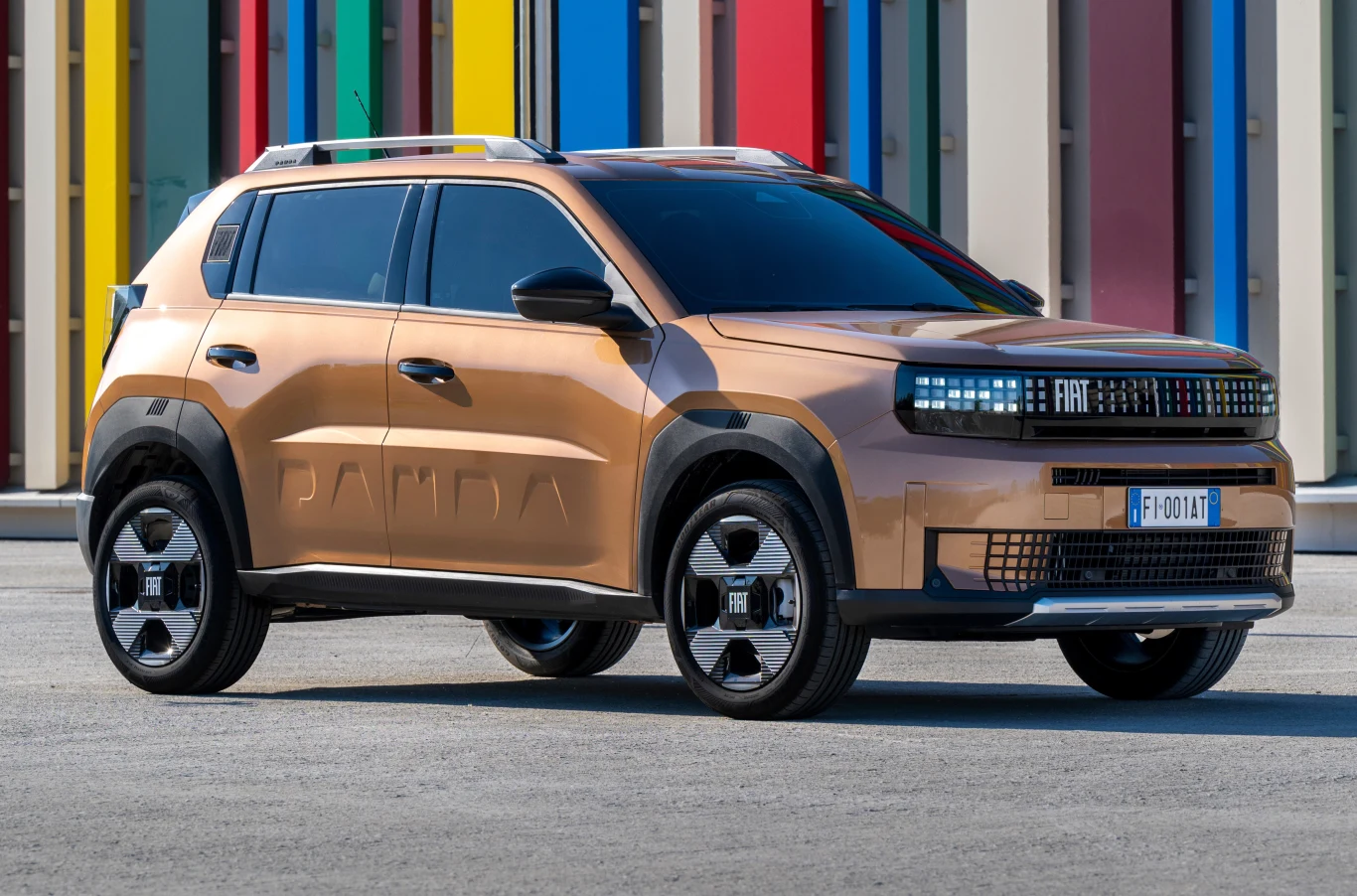 Systemy bezpieczeństwa podnoszą cenę auta. Można tego uniknąć. Nowoczesny, pomarańczowy samochód typu SUV marki Fiat, model Panda, stoi na szarym asfalcie przed tłem kolorowych, pionowych paneli.