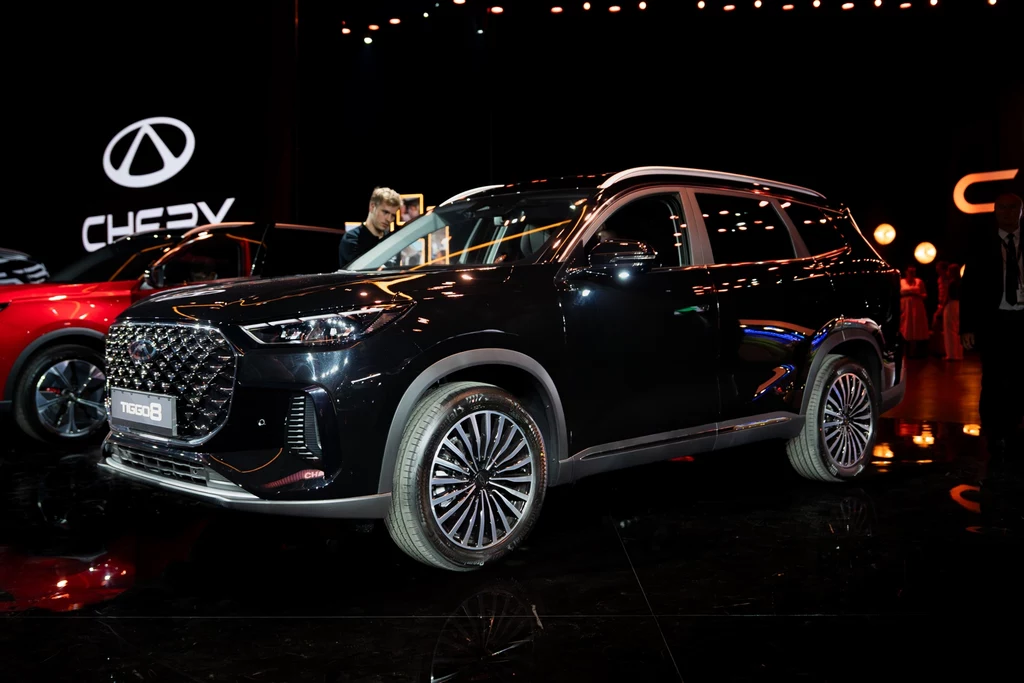 CHERY TIGGO 8 idealny SUV dla dużej rodziny Czarny SUV marki Chery Tiggo 8 prezentowany podczas wydarzenia motoryzacyjnego, nowoczesna sylwetka pojazdu z efektownymi felgami na tle ciemnej, podświetlanej hali wystawowej.