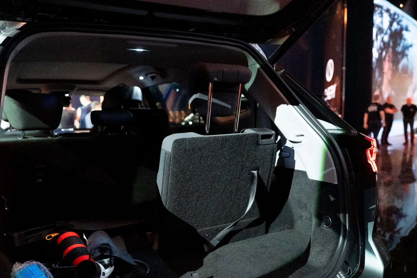 Widok wnętrza bagażnika samochodu z otwartą klapą, złożone tylne siedzenie, na podłodze leży torba lub sprzęt fotograficzny, w tle rozmazane sylwetki osób oraz oświetlone wnętrze budynku.