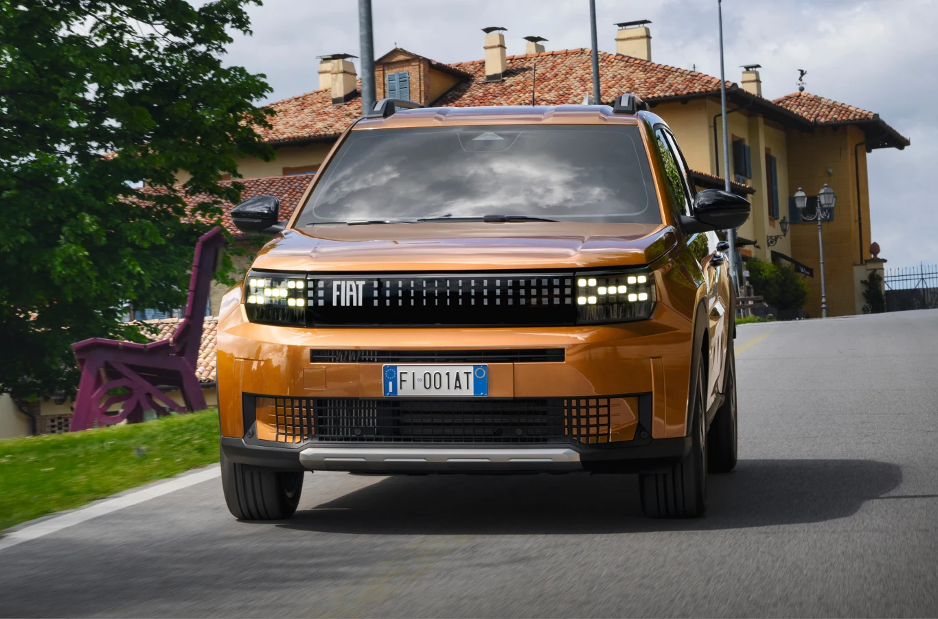 Nowoczesny, pomarańczowy samochód marki Fiat jadący po asfaltowej drodze w otoczeniu budynków z cegły i zieleni, na włoskiej rejestracji.